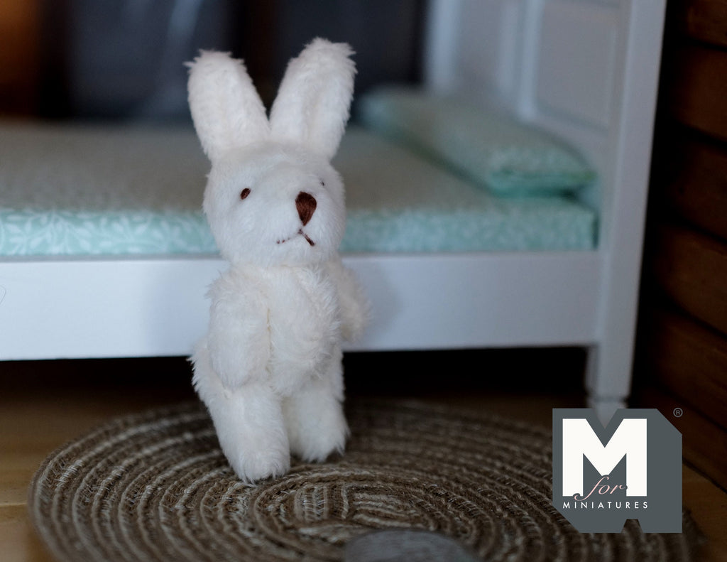 1:12 scale miniature plush bunny doll in cream with long ears, standing on a woven rug beside a bed