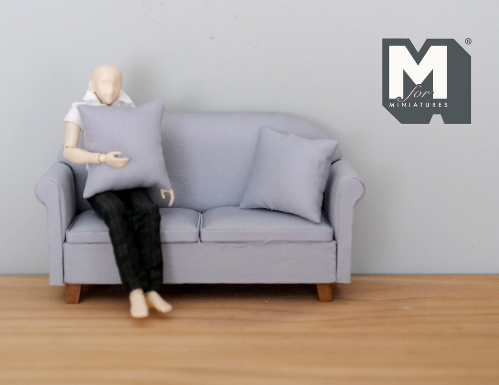 1:12 scale miniature gray fabric 2 seat sofa with pillows and wood legs, shown with a seated figure for scale