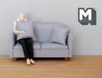 1:12 scale miniature gray fabric 2 seat sofa with pillows and wood legs, shown with a seated figure for scale