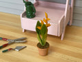 1:12 scale miniature clay pot with yellow flowers and long green leaves, on a wood floor beside garden tools