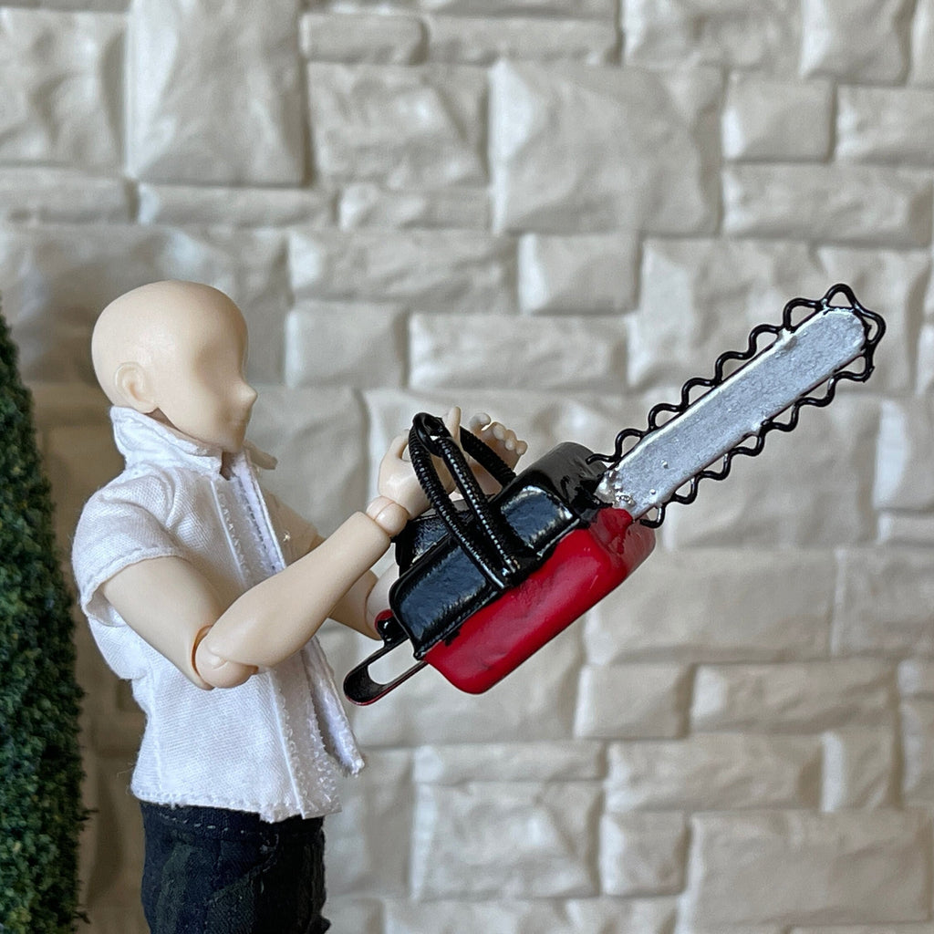 1:12 scale miniature metal chainsaw in red and black with handle and silver bar, held by an artist figure by a stone wall