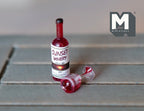 1:12 scale miniature spilled wine set with a red wine bottle and tipped glass, shown on a gray wood table