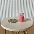 1:12 scale miniature real wax candle in a glass jar with lid, red, shown on a round white table beside a coin