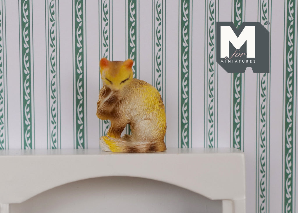 1:12 scale miniature ginger tabby cat, sitting and grooming with raised paw, shown against green striped wallpaper