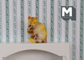 1:12 scale miniature ginger tabby cat, sitting and grooming with raised paw, shown against green striped wallpaper