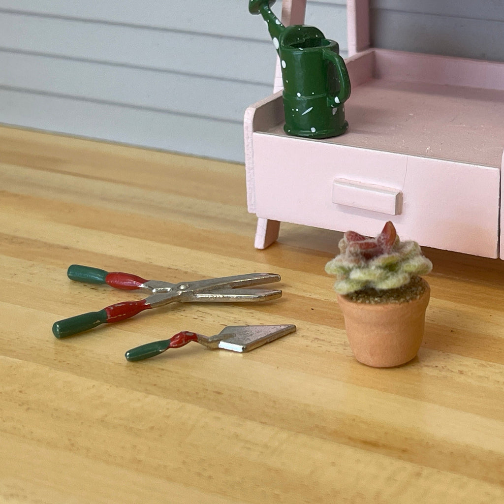 1:12 scale miniature flowering cactus in a clay pot with a pink flower, shown on a wood floor beside garden tools