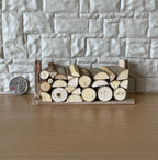 1:12 scale miniature wood log pile on base with stacked split logs, shown against a stone wall beside a coin
