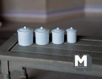1:12 scale miniature kitchen canisters with lids in white, set of 4 graduated sizes, shown in a row on a gray table