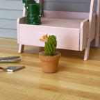 1:12 scale miniature potted spiky cactus with orange blooms in a short clay pot, shown on a wood floor by a pink bench