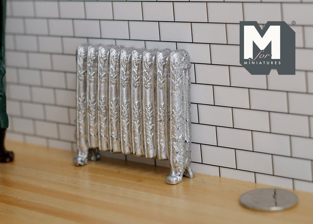 1:12 scale miniature antique style column radiator in silver with ornate feet, against a white tile wall beside a coin