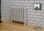 1:12 scale miniature antique style column radiator in silver with ornate feet, against a white tile wall beside a coin