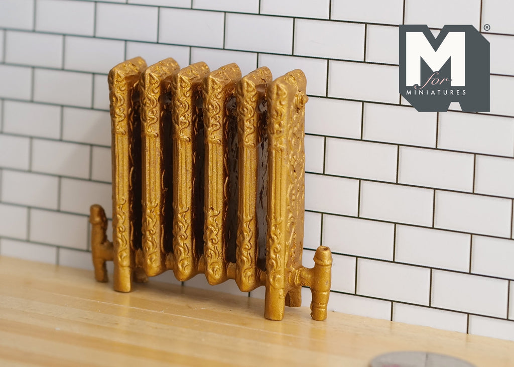 1:12 scale miniature gold antique style column radiator with feet and ornate relief, against a white tile wall