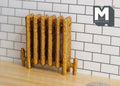 1:12 scale miniature gold antique style column radiator with feet and ornate relief, against a white tile wall