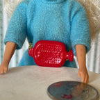 1:12 scale miniature cast resin hot water bottle in red with ribbed texture, held by a doll and shown beside a coin