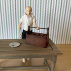 1:12 scale miniature wood garden tool caddy in brown with a top handle, shown on a table beside an artist figure