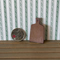 1:12 scale miniature dark brown wood cutting board with handle, shown leaning on a striped wall beside a coin