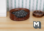 1:12 scale miniature wicker style pet bed in brown with black floral cushion, shown on a wood floor beside a bowl