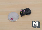 1:12 scale miniature curled up resting cat figure in black with yellow eyes, shown on a wood surface beside a coin