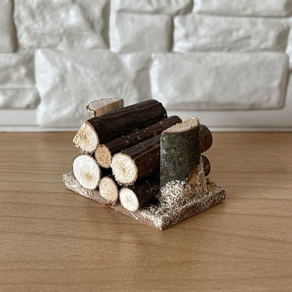 1:12 scale miniature wood log pile on base, stacked cut logs with bark, shown against a white stone wall
