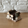1:12 scale miniature wood log pile on base, stacked cut logs with bark, shown against a white stone wall