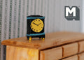 1:12 scale miniature black tabletop clock with yellow dial, blue trim and gold feet, non working, on a wood dresser