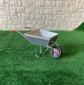 1:12 scale miniature silver metal wheelbarrow with red wheel and grips, shown on green grass by a stone wall