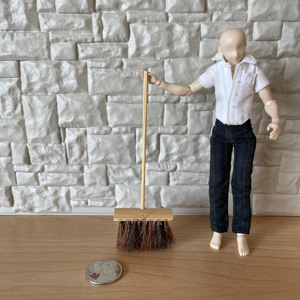 1:12 scale miniature yard brush with wood handle and coarse brown bristles, held by a figure by a stone wall, coin for scale