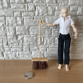 1:12 scale miniature yard brush with wood handle and coarse brown bristles, held by a figure by a stone wall, coin for scale