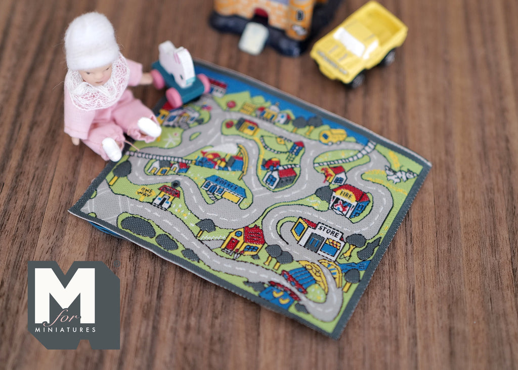 1:12 scale miniature town road play mat with printed streets and houses, shown on a wood floor with a baby doll and toy cars