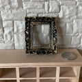 1:12 scale miniature ornate rectangular picture frame in black and gold, on a wood shelf beside a coin for scale