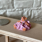 1:12 scale miniature bear figurine in a pink ruffled dress, sitting, shown on a wood shelf beside a coin for scale