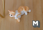 1:12 scale miniature lying cat in ginger and white, crouching pose, shown on a wood floor