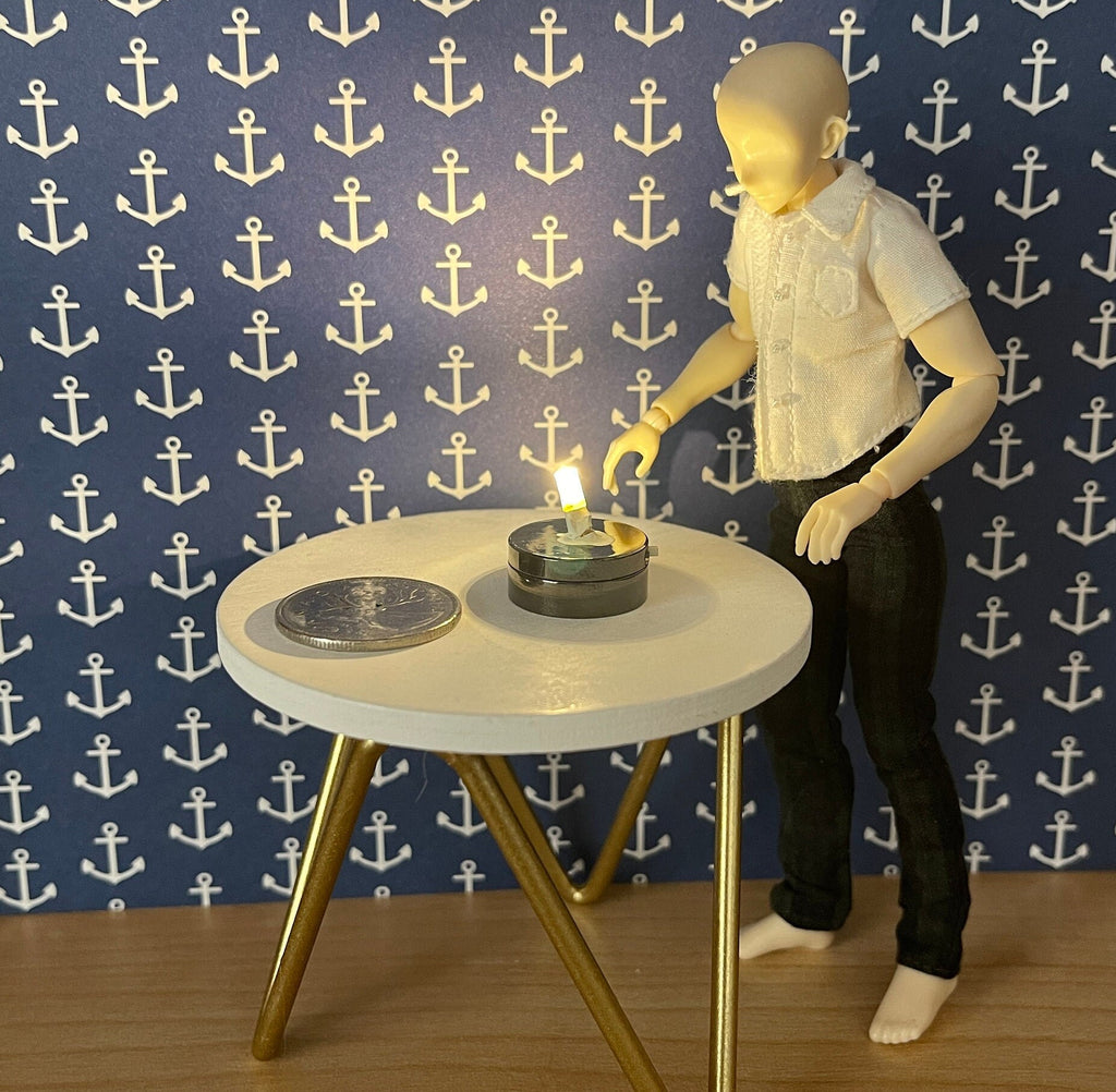 1:12 scale miniature LED light bulb with black base, battery powered, shown lit on a round table beside a figure