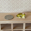 1:12 scale miniature lying tabby cat in gray and tan, curled with head up, shown on a wood shelf beside a coin