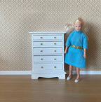 1:12 scale miniature wood tall dresser in white with 5 drawers and gold knobs, shown beside a doll in a blue dress
