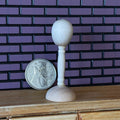 1:12 scale miniature white mannequin head on tall round stand, shown beside a coin for scale against a purple brick wall