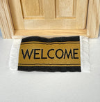 1:12 scale miniature welcome doormat in black and tan with white fringe, shown at a wood front door