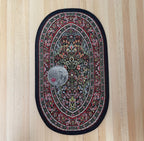 1:12 scale miniature oval area rug with floral medallion in red, green and black, shown on a wood floor with a coin