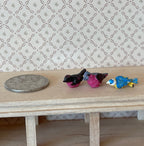 1:12 scale miniature small birds in assorted colors, set of 3, black, pink and blue, on a wood shelf beside a coin