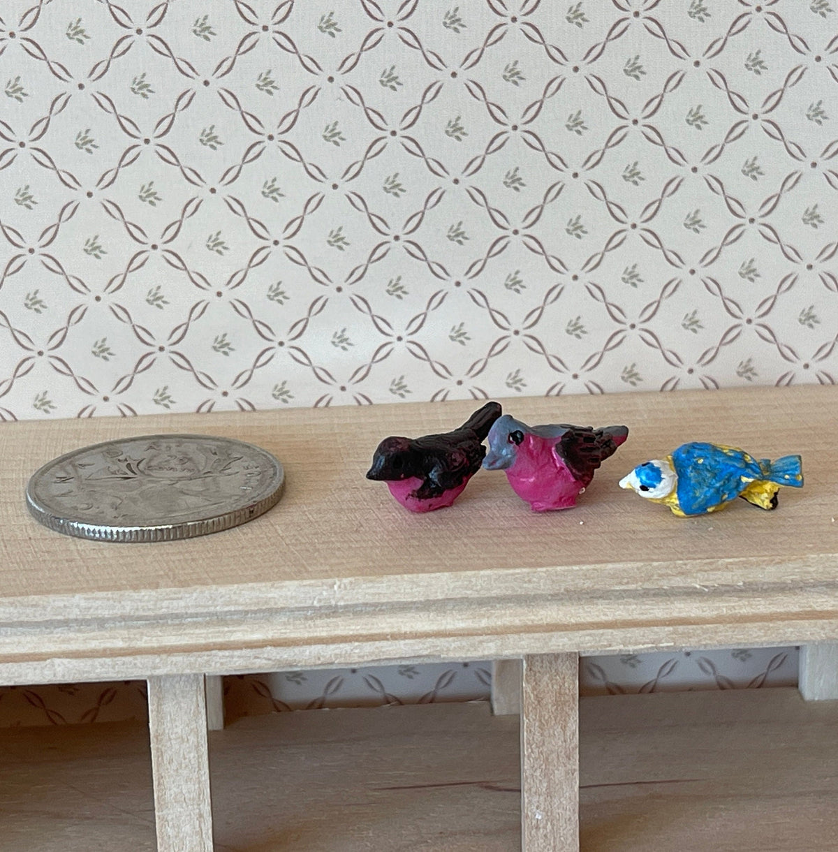 1:12 scale miniature small birds in assorted colors, set of 3, black, pink and blue, on a wood shelf beside a coin