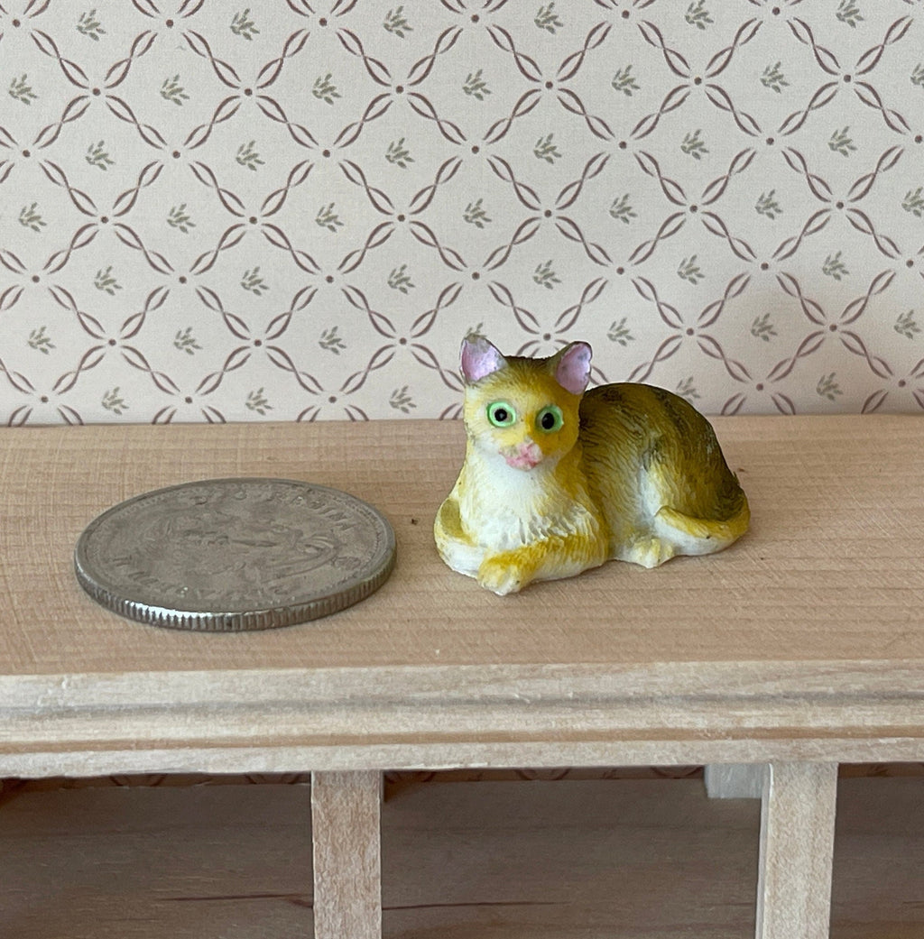 1:12 scale miniature lying tabby cat in tan with green eyes, style A, shown on a wood shelf beside a coin