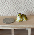 1:12 scale miniature lying tabby cat in tan with green eyes, style A, shown on a wood shelf beside a coin