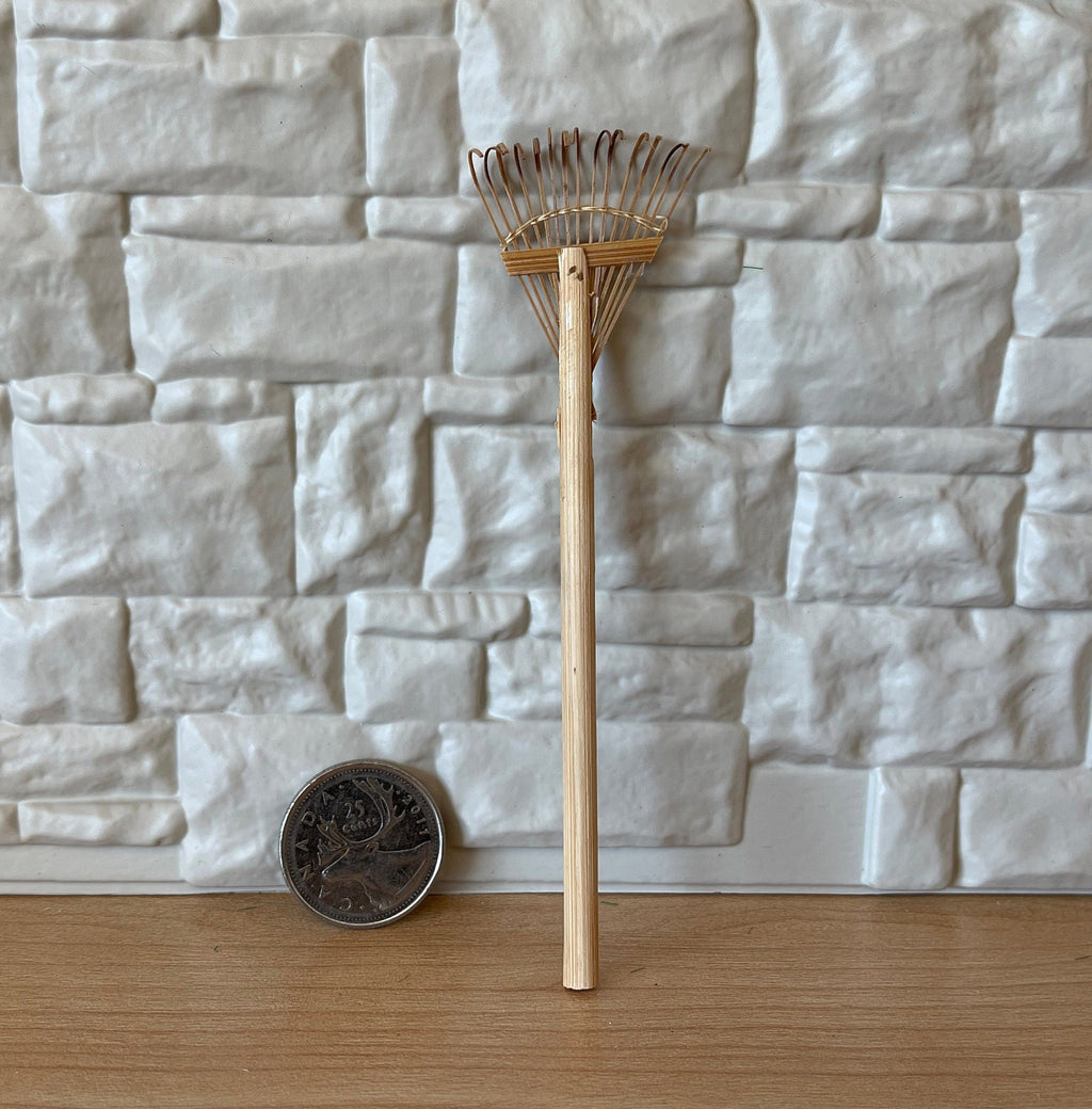 1:12 scale miniature leaf rake with wood handle and fan shaped metal tines, leaning on a stone wall beside a coin for scale