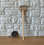 1:12 scale miniature leaf rake with wood handle and fan shaped metal tines, leaning on a stone wall beside a coin for scale
