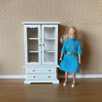 1:12 scale miniature wood dish cabinet in white with glass doors and drawers, shown beside a doll in a blue dress