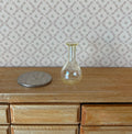 1:12 scale miniature clear glass decanter vase with round base and flared neck, shown on a wood dresser beside a coin
