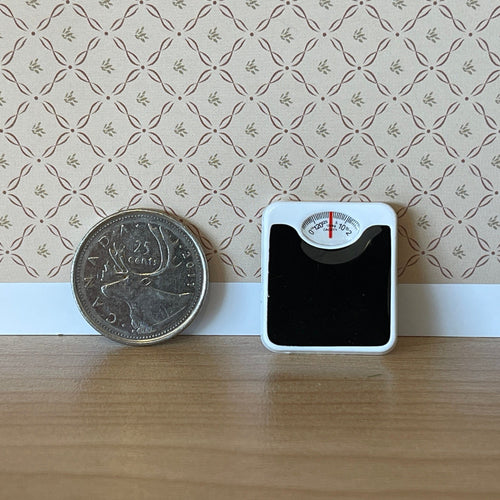 1:12 scale miniature bathroom scale with round dial, black platform with white trim, standing beside a coin for scale
