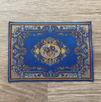 1:12 scale miniature area rug in royal blue with a floral medallion and ornate border, shown flat on a wood floor