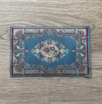 1:12 scale miniature area rug in blue with a floral medallion and red patterned border, shown flat on a wood floor