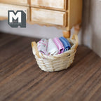 1:12 scale miniature bamboo rope basket with folded linens in pink, blue and white fixed inside, on a wood floor